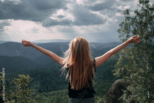 Young blond girl with hands up enjoing view of the evergreen taiga forest hills on rainy day. Stolby Nature Reserve in Krasnoyarsk, Russia.