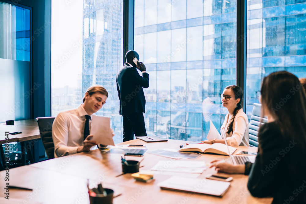 African American male boss calling to corporate partner using office internet connection on smartphone while confident employees analyzing report documents, formally dressed businessman phoning African American male boss calling to corporate partner using office internet connection on smartphone while confident employees analyzing report documents, formally dressed businessman phoning