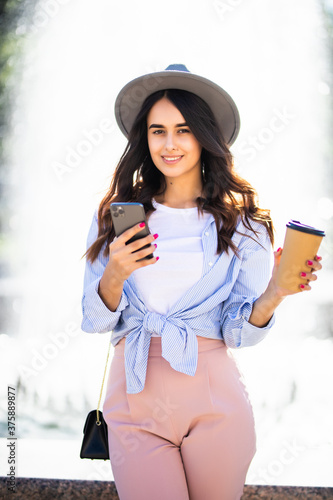 Cheerful girl in the street drinking morning coffee and use her smartphone. Young woman using phone on the street. Woman texting.