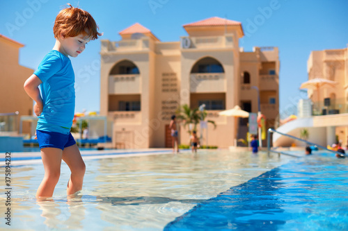 toddler boy in ultraviolet protective swimsuit walking in shallow water in the pool during summer vacation