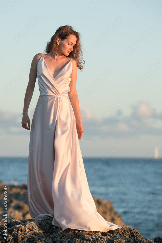 Woman in silky flowing dress. Graceful caucasian woman in silk pink flowing dress on the rocky shore against unfocused sea Woman in silky flowing dress. Graceful caucasian woman in silk pink flowing dress on the rocky shore against unfocused sea