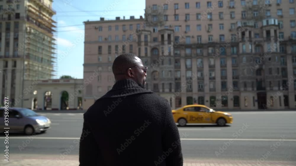 An African-American man walks on a city street against the background of a highway with cars and city houses. He is wearing dark glasses.