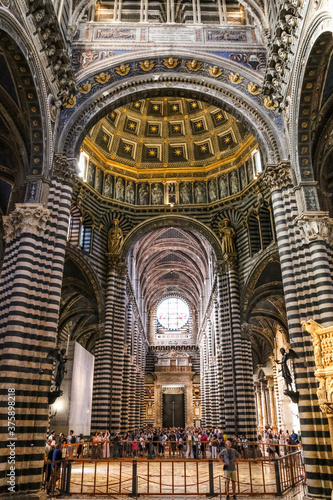 Magnificent view of the central nave with black and white marble stripes on walls and columns inside the Duomo di Siena. Visitors admire the marble mosaic floor and the roof of the hexagonal dome.