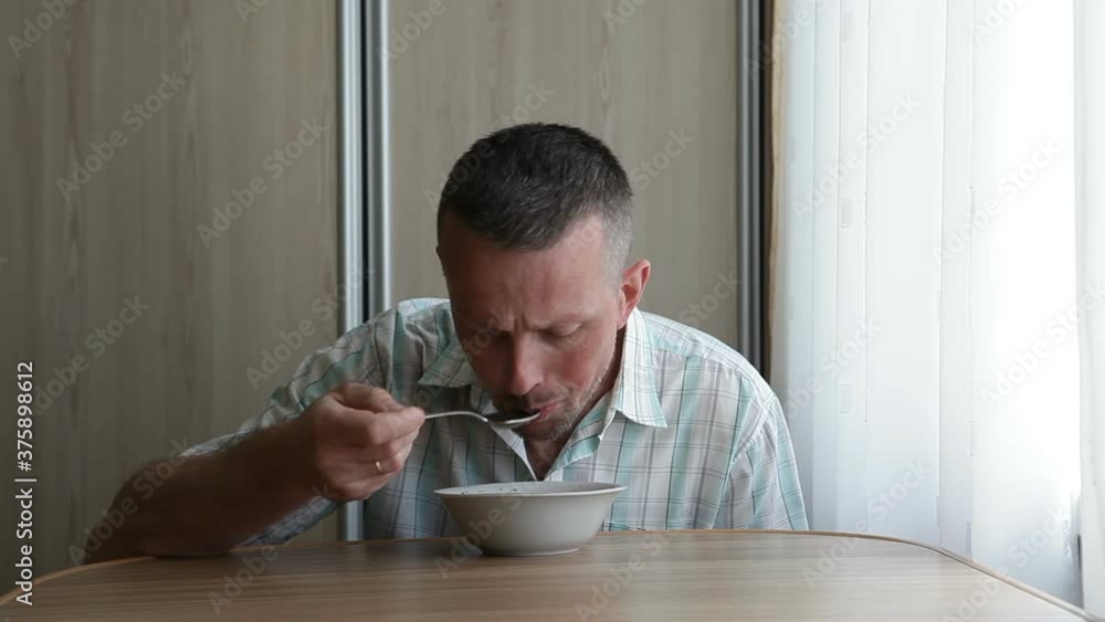 A man sits at a table near the window and dines. The man is eating soup. A man sits at a table near the window and dines. The man is eating soup.