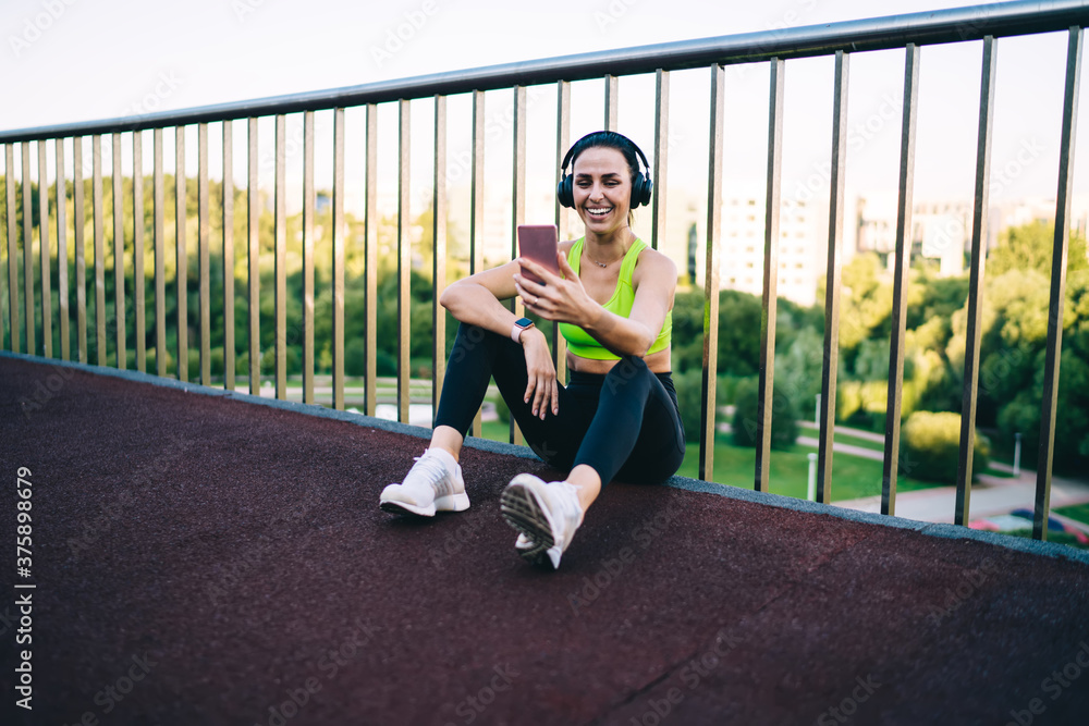 Prosperous female fitness influencer laughing making vlog video on smartphone camera after training outdoors, cheerful woman blogger posing for selfie on smartphone listening music in headphones