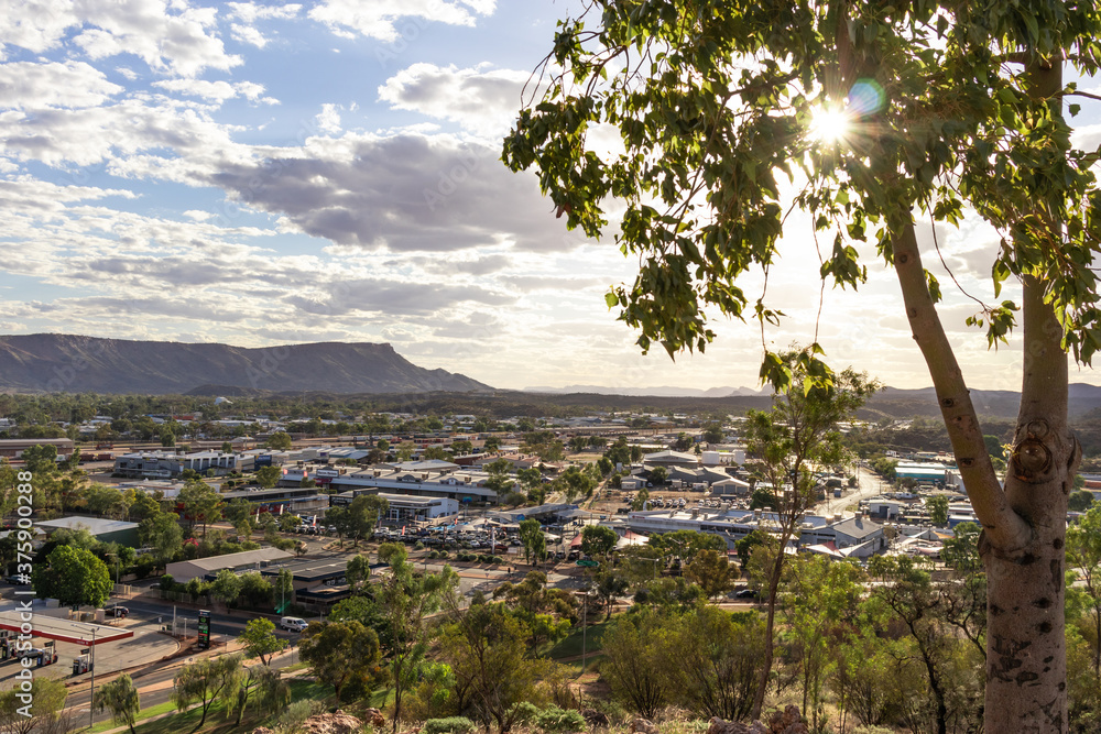 Views of Alice springs township from Anzac hill. Mountains MacDonnell ranges at background. Sun shining among the tree leaves at sunset time. Cloudy sky. Alice Springs, Australia