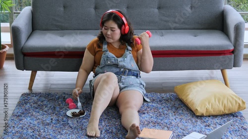 The overweight young woman sitting on the carpet in the living room wearing red headphones, eat chocolate cake while exercise. Look chill and have fun at home.