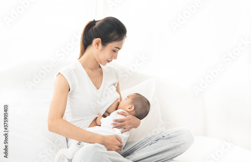 Young asian mother feeding breast her baby on bed at home in white room. Asia mom holding her baby and looking to her child. Woman and new born relax in a white bedroom.