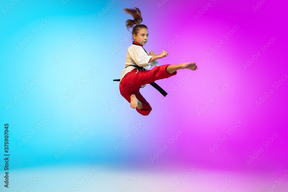 In jump. Karate, taekwondo girl with black belt isolated on gradient background in neon light. Little caucasian model, sport kid training in motion and action. Sport, movement, childhood concept.