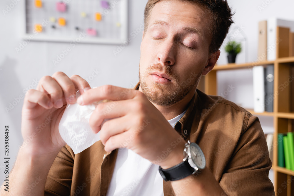 Selective focus of sick businessman with closed eyes holding napkin in office Selective focus of sick businessman with closed eyes holding napkin in office