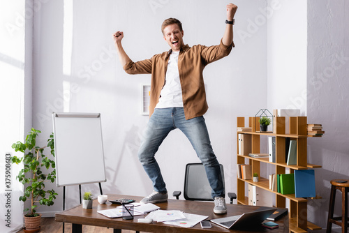 Schilderij op canvas Businessman showing yeah gesture while standing near gadgets and papers on offic