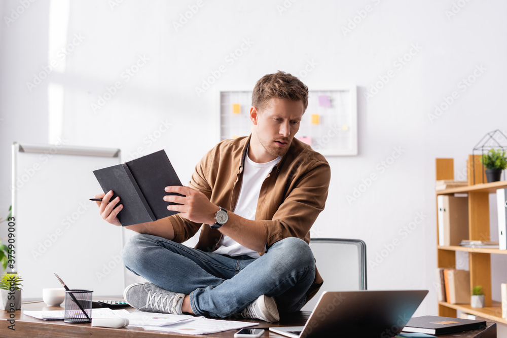 Selective focus of businessman holding pen and notebook while sitting near laptop and papers on office table Selective focus of businessman holding pen and notebook while sitting near laptop and papers on office table
