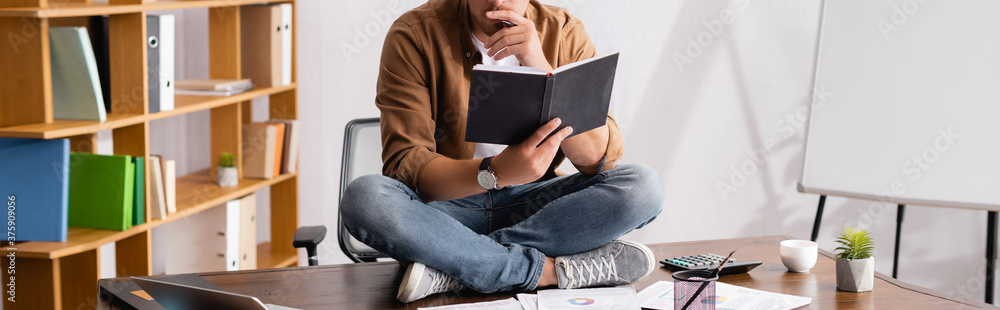 Fototapeta premium Panoramic crop of businessman holding notebook while sitting on table in office
