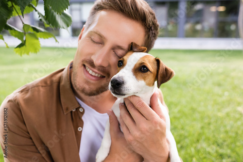 young man with closed eyes holding white jack russell terrier dog with brown spots on head