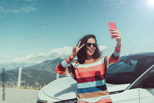 Happy young woman making a self-portrait. She is supported on white car. She is wearing colorful scratch jersey. She is in a mountain road with mountain landscape with blue sky on back