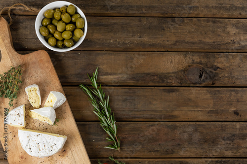 View of a wooden cutting board with cheese on a textured wooden surface with thyme and bowl of olive
