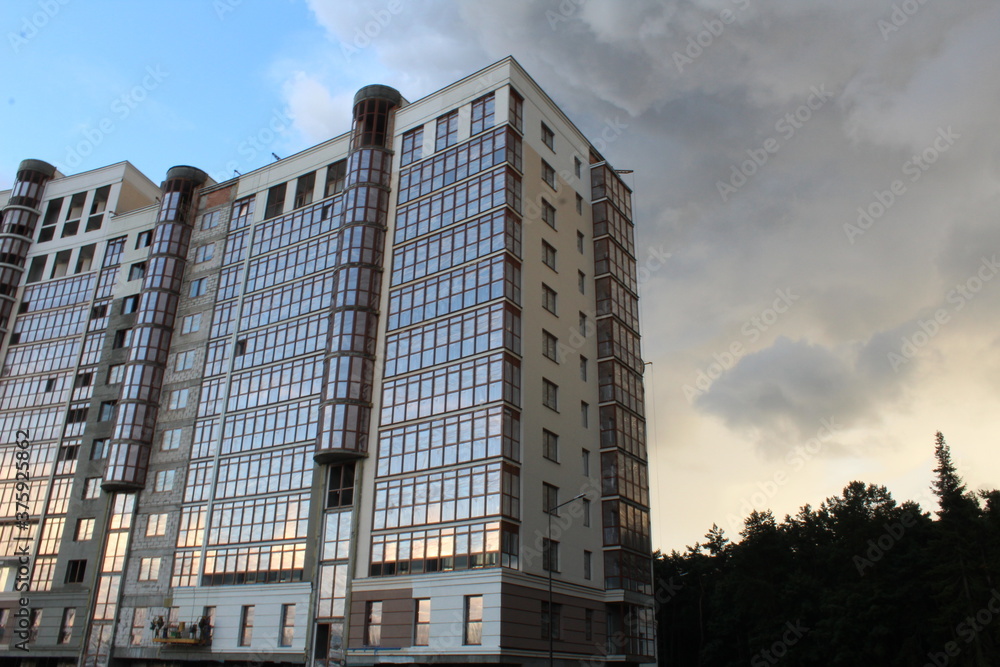 Fototapeta premium the building is a modern residential building complex with a glass facade during construction on the background of a blue sky with clouds illuminated by the sun at sunset