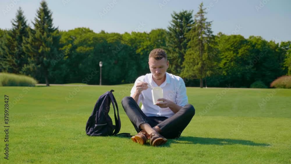 Businessman eats vegetarian lunch with an ecologic package frowning showing dislike. Guy sits in park on green grass eats salad. Wooden sticks. Paper packaging Businessman eats vegetarian lunch with an ecologic package frowning showing dislike. Guy sits in park on green grass eats salad. Wooden sticks. Paper packaging