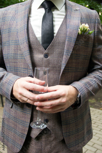 A man in a three-piece suit holds a glass. To close. A man's hand holds a glass of white wine. The groom's suit