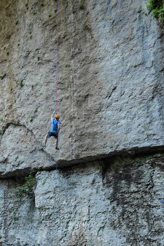 A rock climber in full uniform is going down a gray rock. The athlete climbs the rock with the help of the ropes.