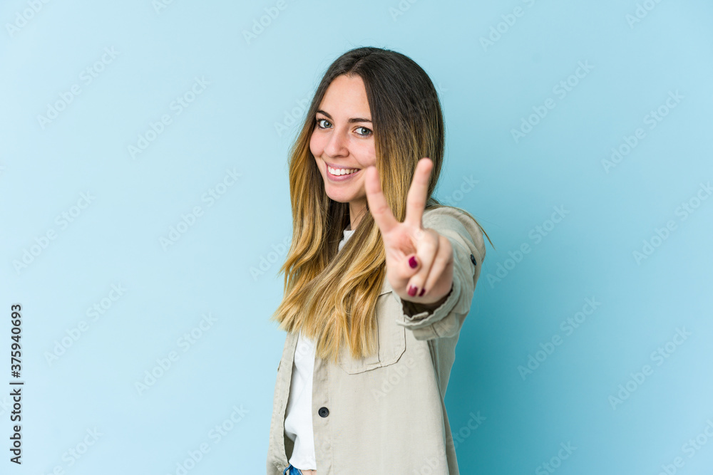 Young caucasian woman isolated on blue background joyful and carefree showing a peace symbol with fingers. Young caucasian woman isolated on blue background joyful and carefree showing a peace symbol with fingers.