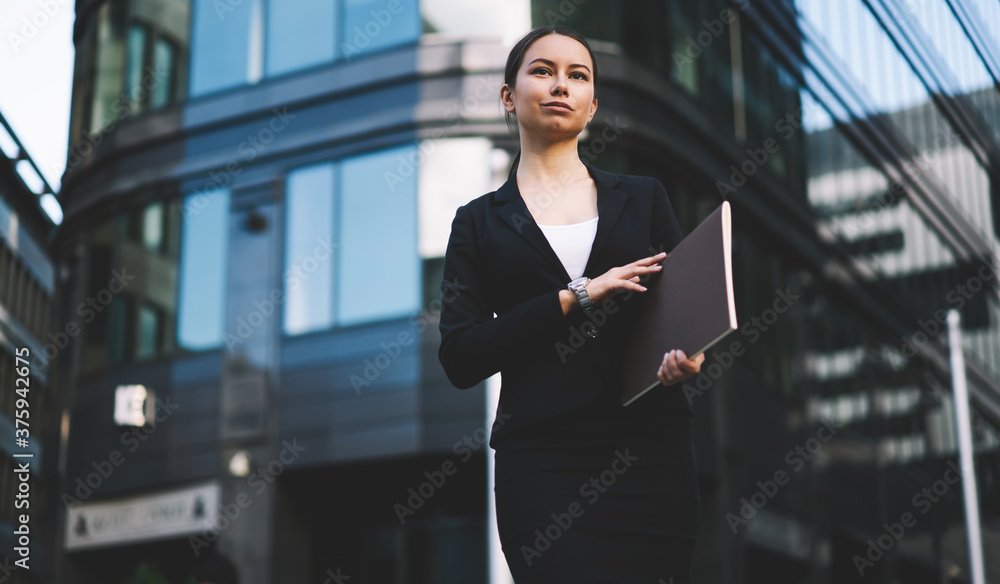 Good looking woman with folder in hands looking away near entrance of modern building