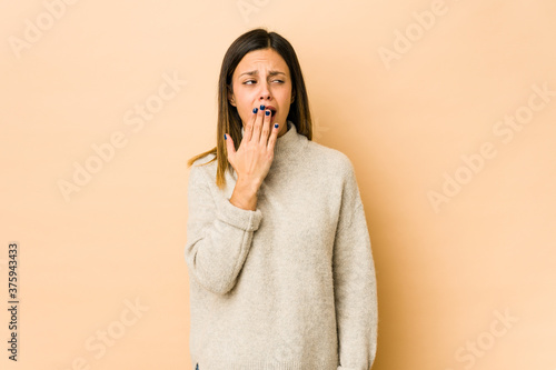 Young woman isolated on beige background yawning showing a tired gesture covering mouth with hand.