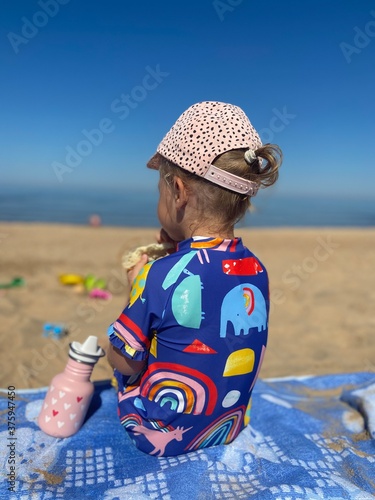 little girl in colorful swimsuit sitting on the blue blanket at the beach on a dummy summer warm day