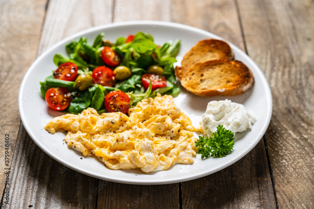 Delicious breakfast - scrambled eggs with fried sausages, cottage cheese and vegetables served on black plate on wooden table
