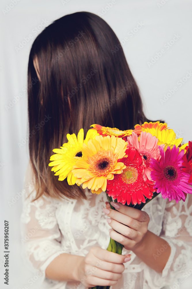 Naklejka premium Beautiful woman in white dress hid her face behind large bouquet of colorful bright gerberas.