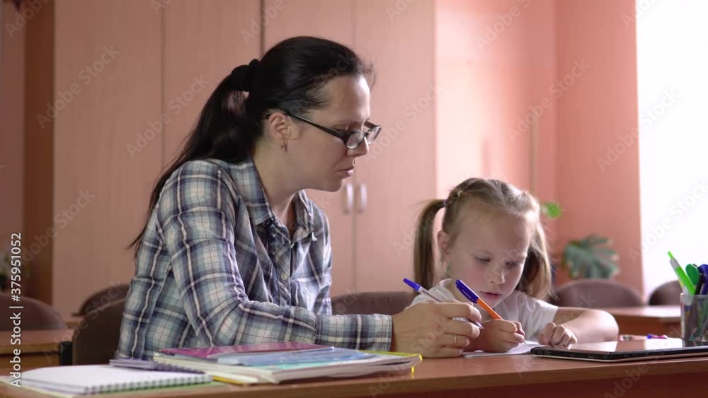Little beautiful girl is studies with the teacher in the school class. Draws in a notebook with a pen. Preschool education.4k Little beautiful girl is studies with the teacher in the school class. Draws in a notebook with a pen. Preschool education.4k