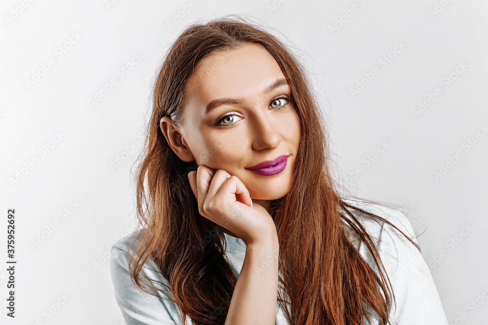 Beautiful sweet brunette girl with long hair with fashion makeup with purple lips on an isolated gray background. The woman holds her hand to her face and looks into the camera. Kind cute model Beautiful sweet brunette girl with long hair with fashion makeup with purple lips on an isolated gray background. The woman holds her hand to her face and looks into the camera. Kind cute model