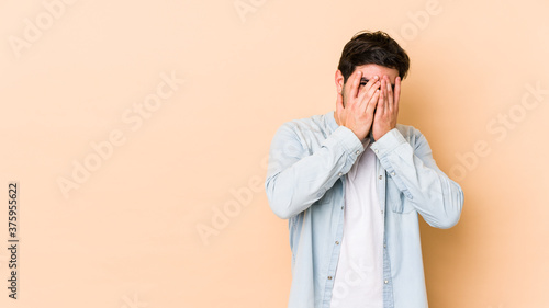 Young man isolated on beige background blink at the camera through fingers, embarrassed covering face.