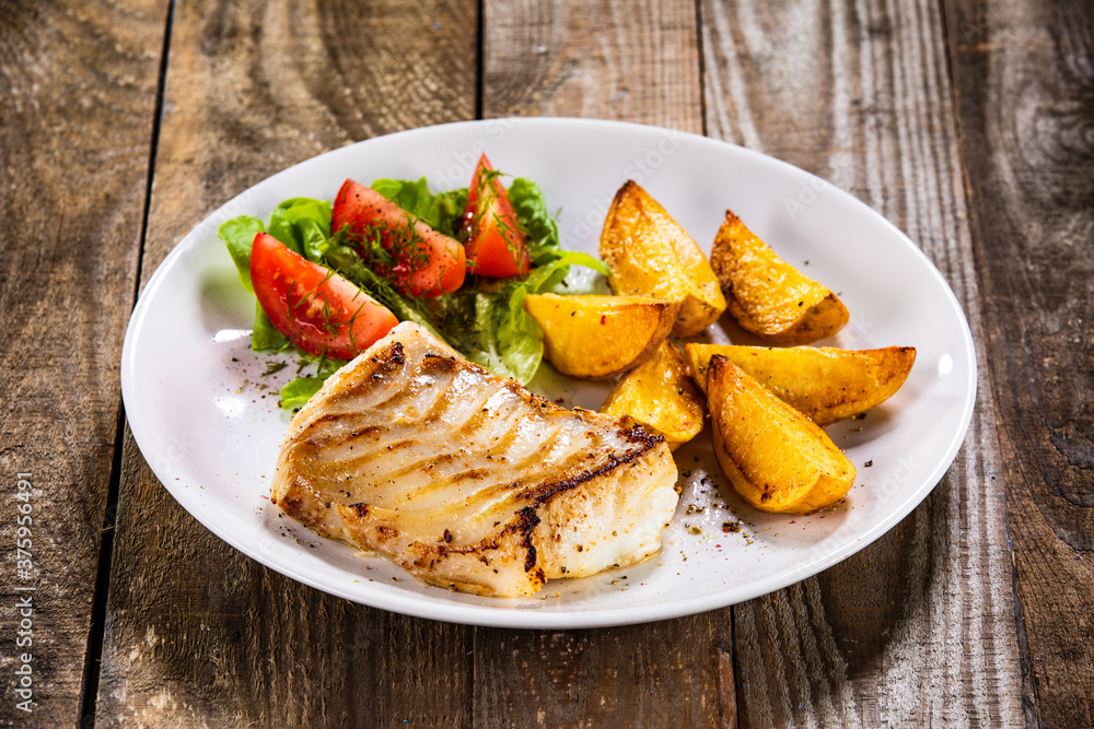 Fish dish - fried cod fillet with potatoes and vegetable salad on wooden table Fish dish - fried cod fillet with potatoes and vegetable salad on wooden table