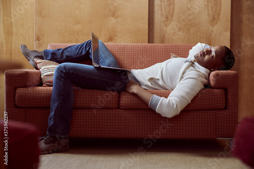 Young man taking a nap on loveseat with his computer on his lap