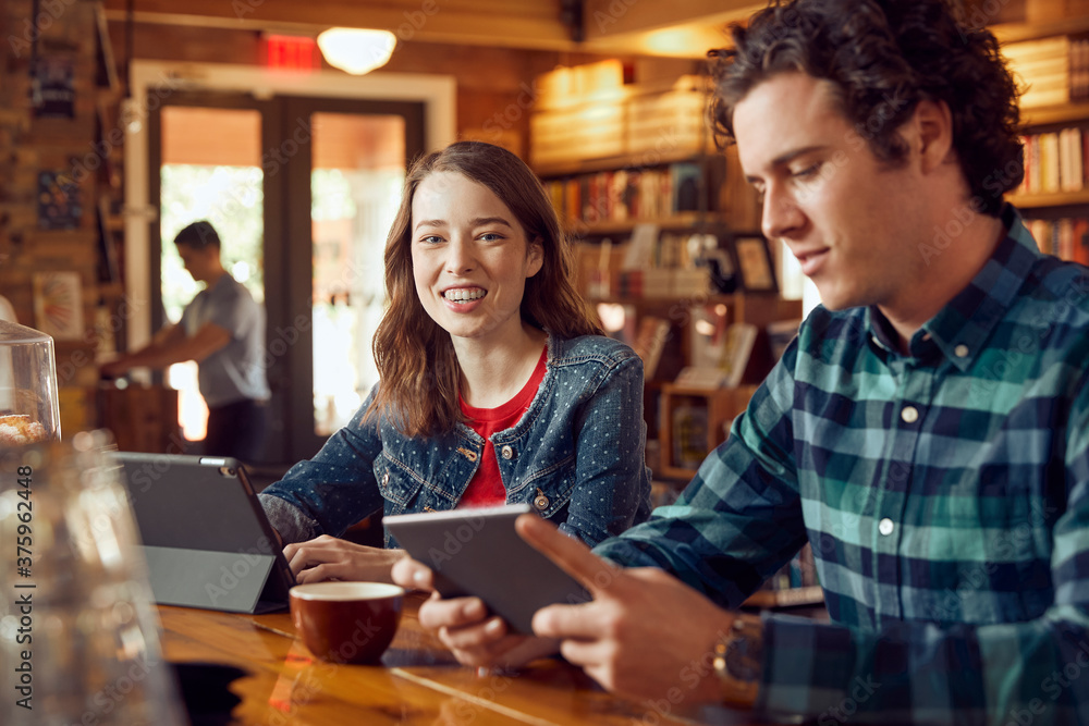 Young man and woman sitting at counter in cafe bookstore using digital tablets , woman looking at camera smiling Young man and woman sitting at counter in cafe bookstore using digital tablets , woman looking at camera smiling