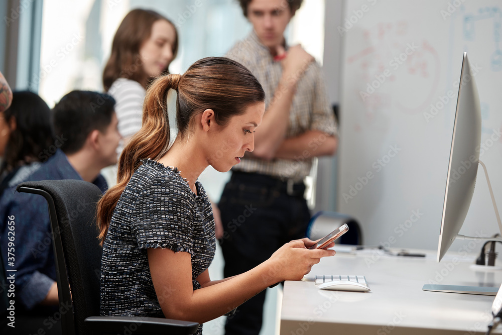 © SuperStock - Young woman using cell phone at computer, co-workers having discussion around dry erase board in background