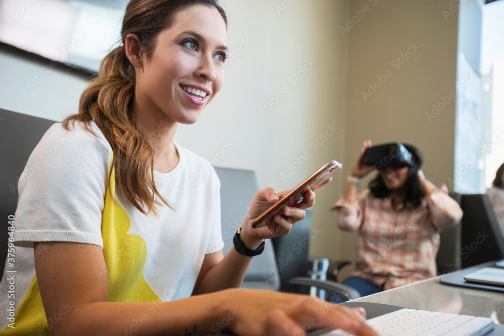 Young woman in conference room working on computer checking mobile phone, with co-worker in background wearing a Virtual reality headset Young woman in conference room working on computer checking mobile phone, with co-worker in background wearing a Virtual reality headset