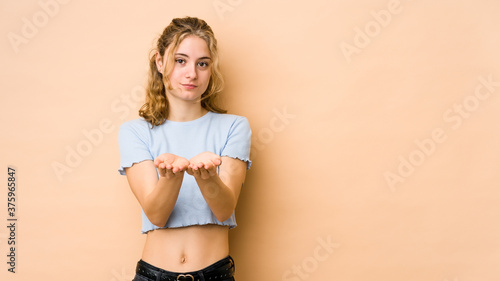 Young caucasian woman isolated on beige background holding something with palms, offering to camera.