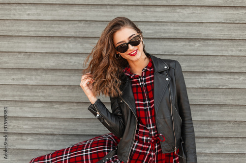Cute stylish joyful young woman with red lipstick with positive smile in sunglasses in fashion leather jacket near vintage wooden wall on street. Happy smiling woman model straightens hair in city.