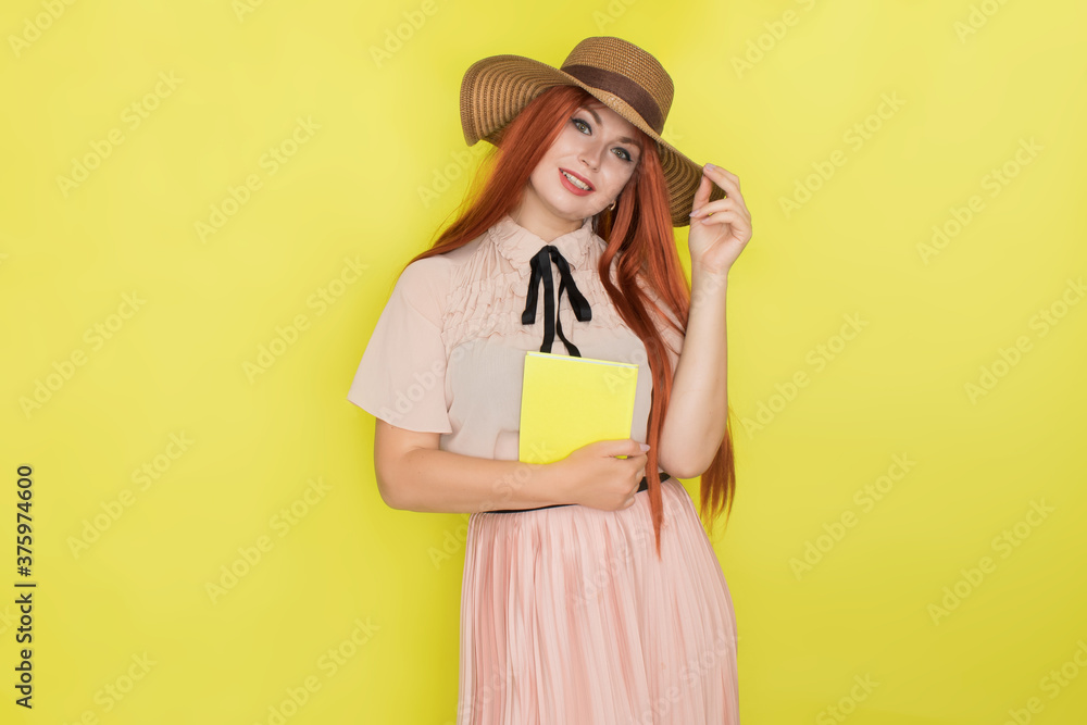 Red haired woman in straw hat over yellow background holding book in her hands Red haired woman in straw hat over yellow background holding book in her hands