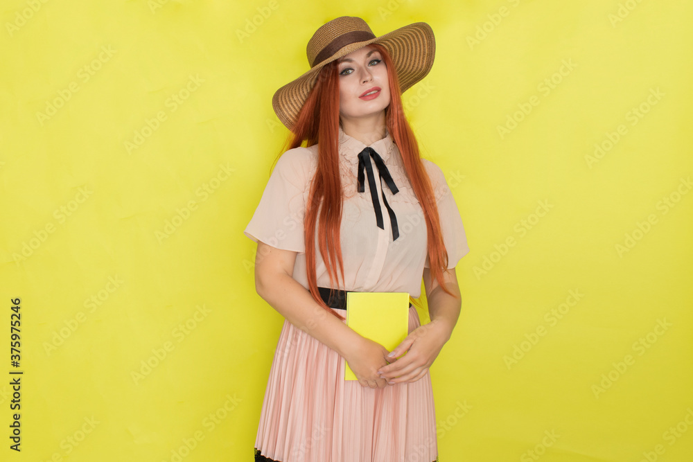 Red haired woman in straw hat over yellow background holding book in her hands Red haired woman in straw hat over yellow background holding book in her hands
