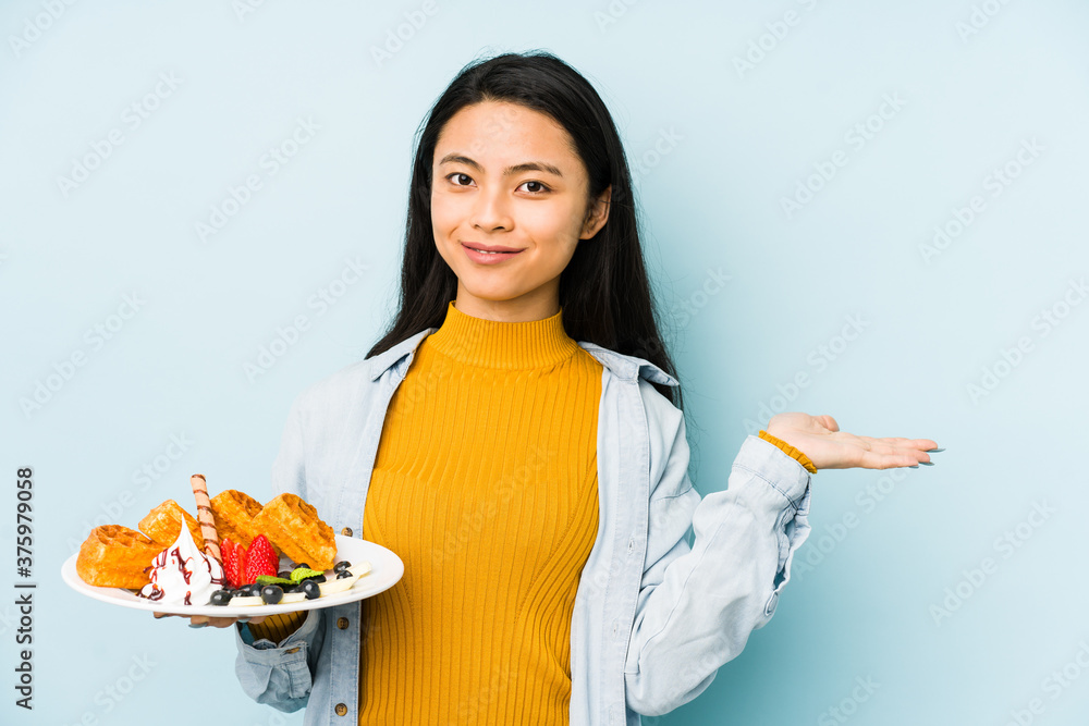 Young chinese woman holding waffle isolated on blue background relaxed thinking about something looking at a copy space. Young chinese woman holding waffle isolated on blue background relaxed thinking about something looking at a copy space.