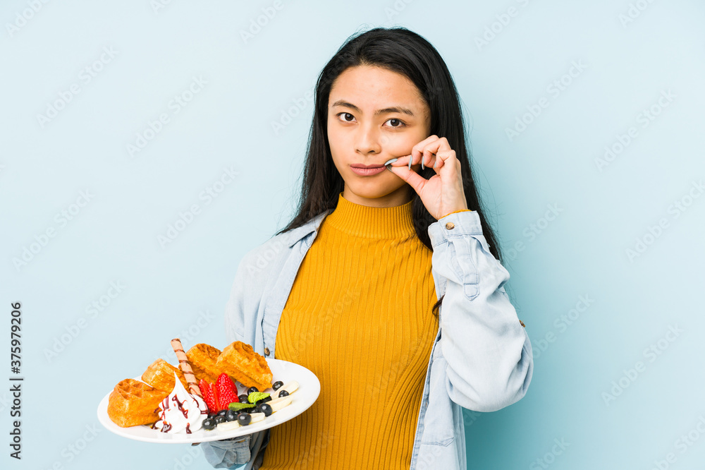 Young chinese woman holding waffle isolated on blue background shrugs shoulders and open eyes confused. Young chinese woman holding waffle isolated on blue background shrugs shoulders and open eyes confused.