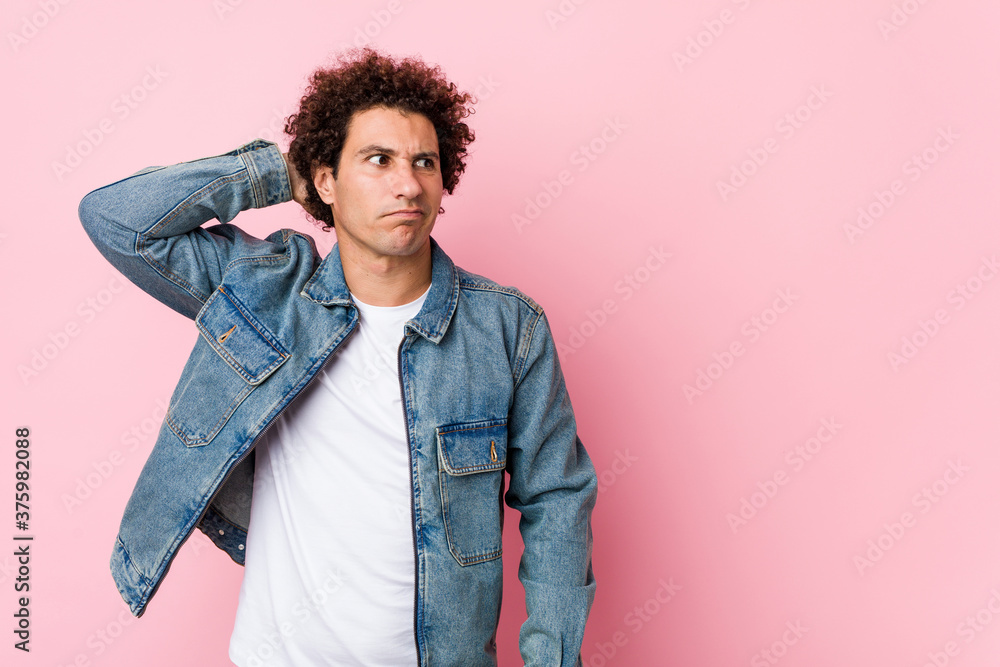 Fototapeta premium Curly mature man wearing a denim jacket against pink background touching back of head, thinking and making a choice.