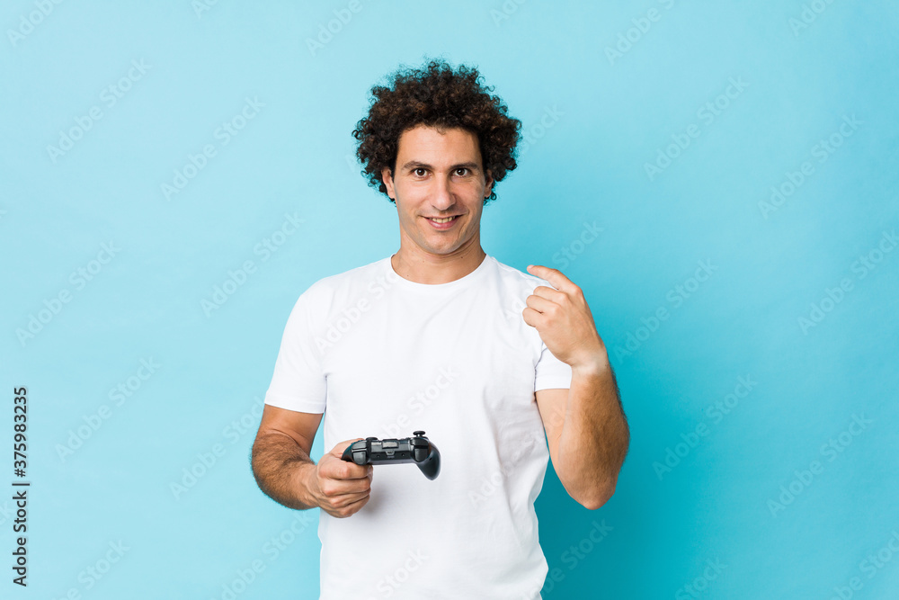 Young caucasian curly man holding a game controller pointing with finger at you as if inviting come closer. Young caucasian curly man holding a game controller pointing with finger at you as if inviting come closer.