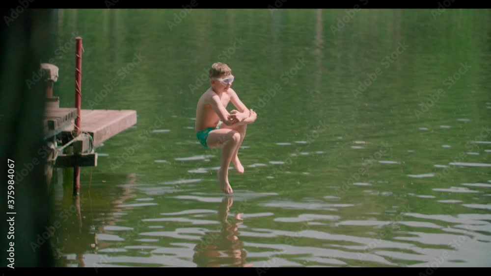 Behind tree pan shot short of a Caucasian kid jumping from the wooden pier into the lake. Behind tree pan shot short of a Caucasian kid jumping from the wooden pier into the lake.