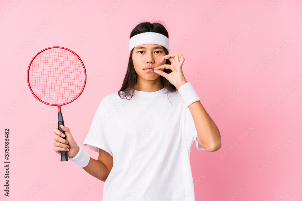 Young chinese woman playing badminton in a pink background with fingers on lips keeping a secret. Young chinese woman playing badminton in a pink background with fingers on lips keeping a secret.