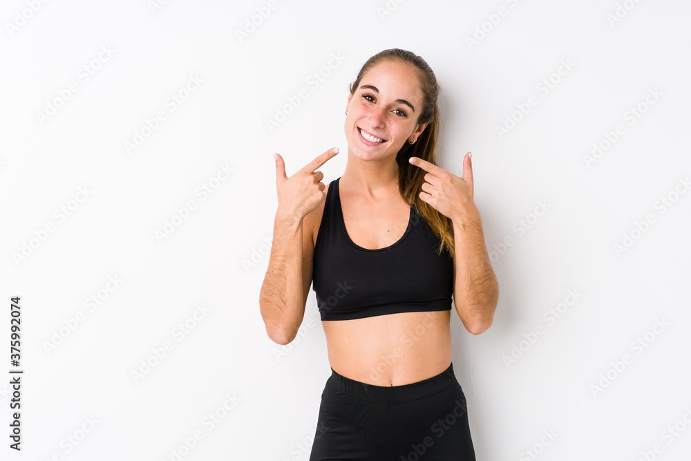 Young caucasian fitness woman posing in a white background smiles, pointing fingers at mouth. Young caucasian fitness woman posing in a white background smiles, pointing fingers at mouth.