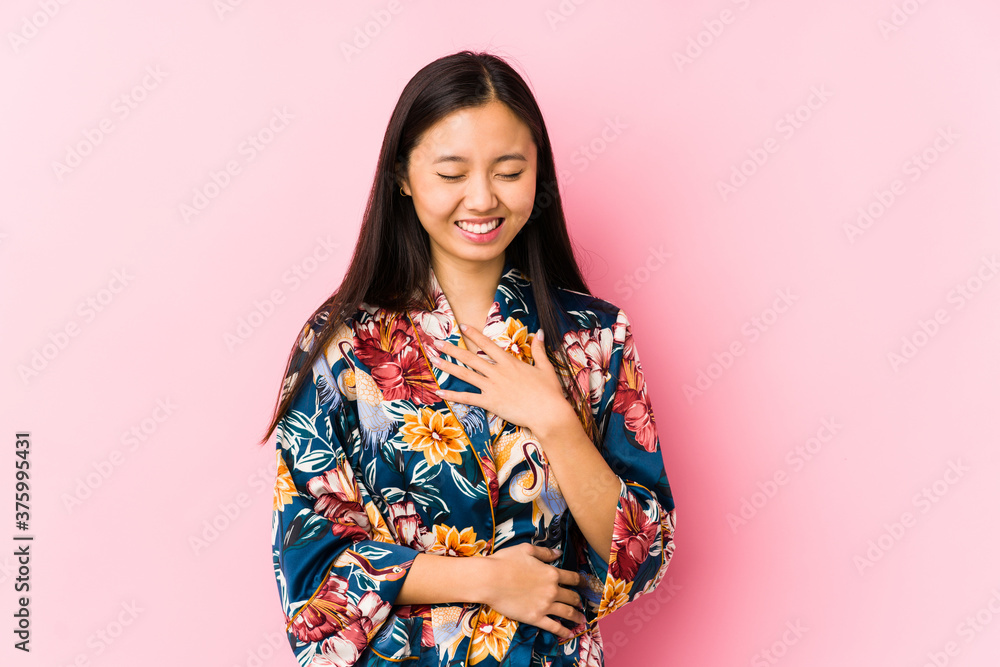 Young chinese woman wearing a kimono pajama isolated laughs happily and has fun keeping hands on stomach. Young chinese woman wearing a kimono pajama isolated laughs happily and has fun keeping hands on stomach.